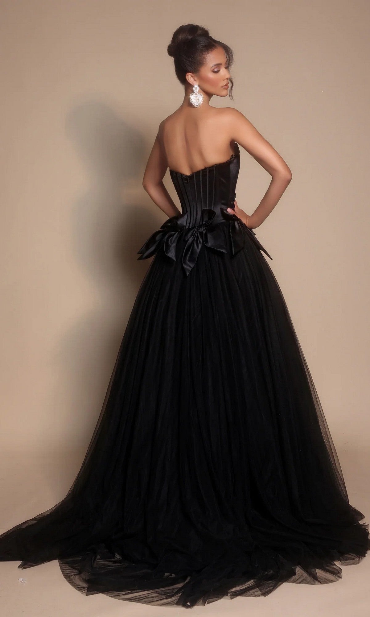 A woman models the Portia and Scarlett PS26102 strapless ball gown, featuring a fitted corset bodice, full tulle skirt, and a large waist bow, photographed from behind against a plain background.